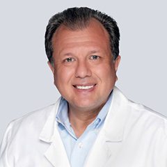 Smiling man in a white lab coat and light blue shirt against a gray background