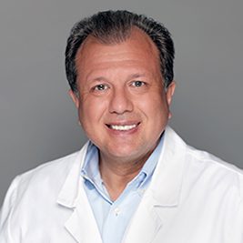 Smiling man in a white lab coat and light blue shirt against a gray background